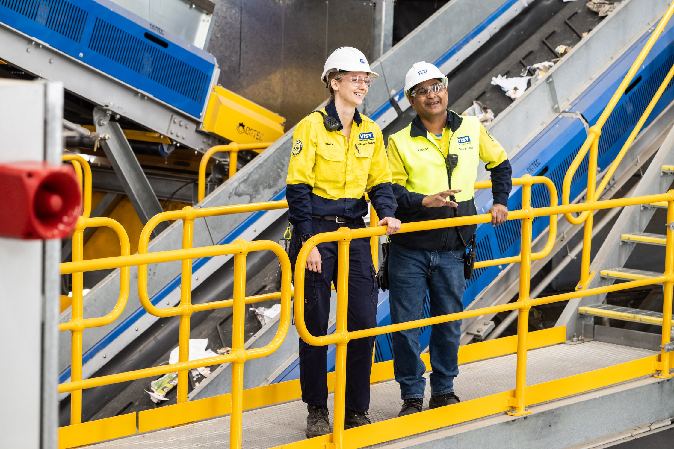 Two people smiling in high visibility safety clothes and hard hats are standing on a walk way in an industrial looking building