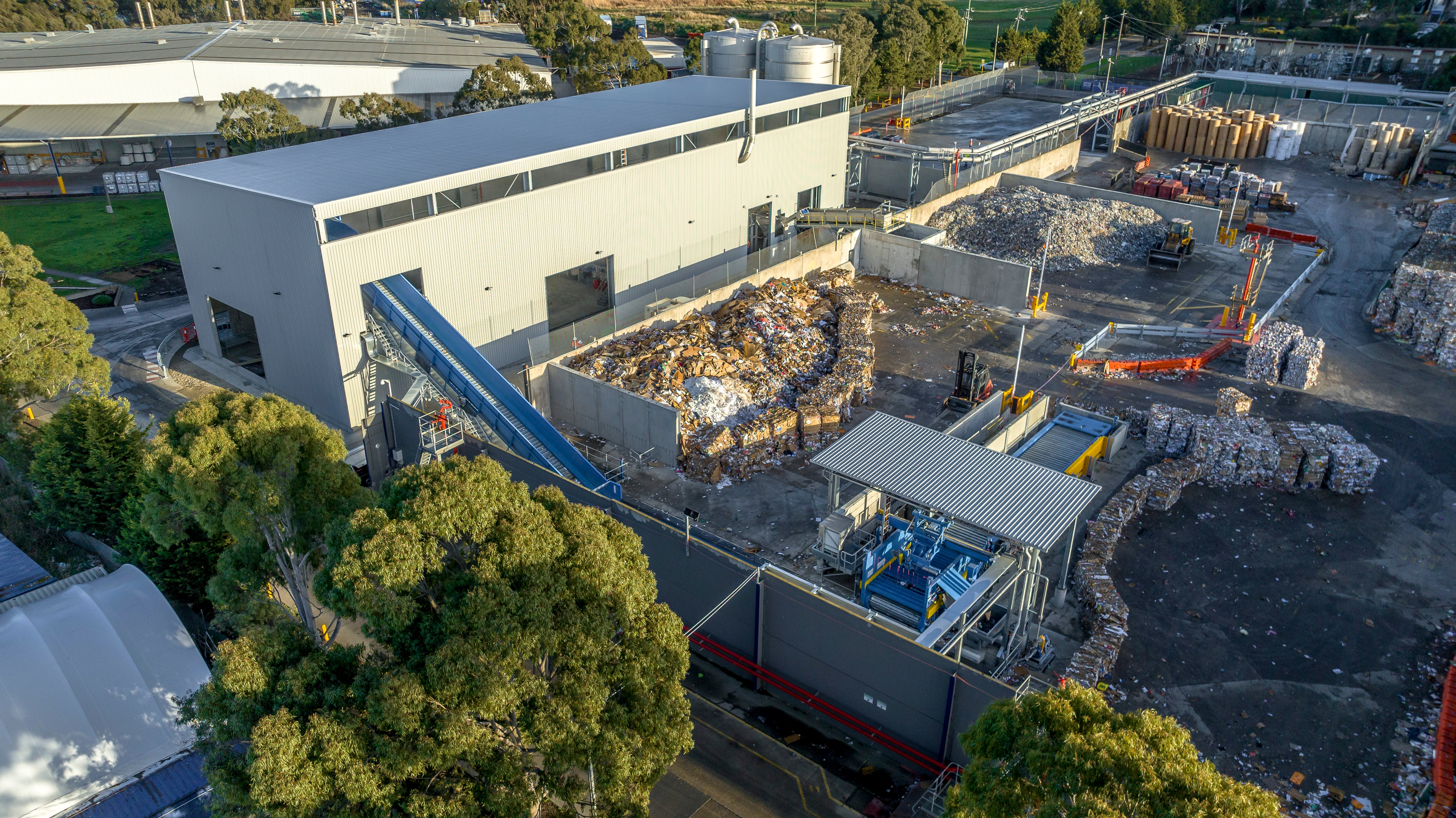 A paper recycling facility, which looks industrial, with a large shed, blue conveyor belt and bales of paper