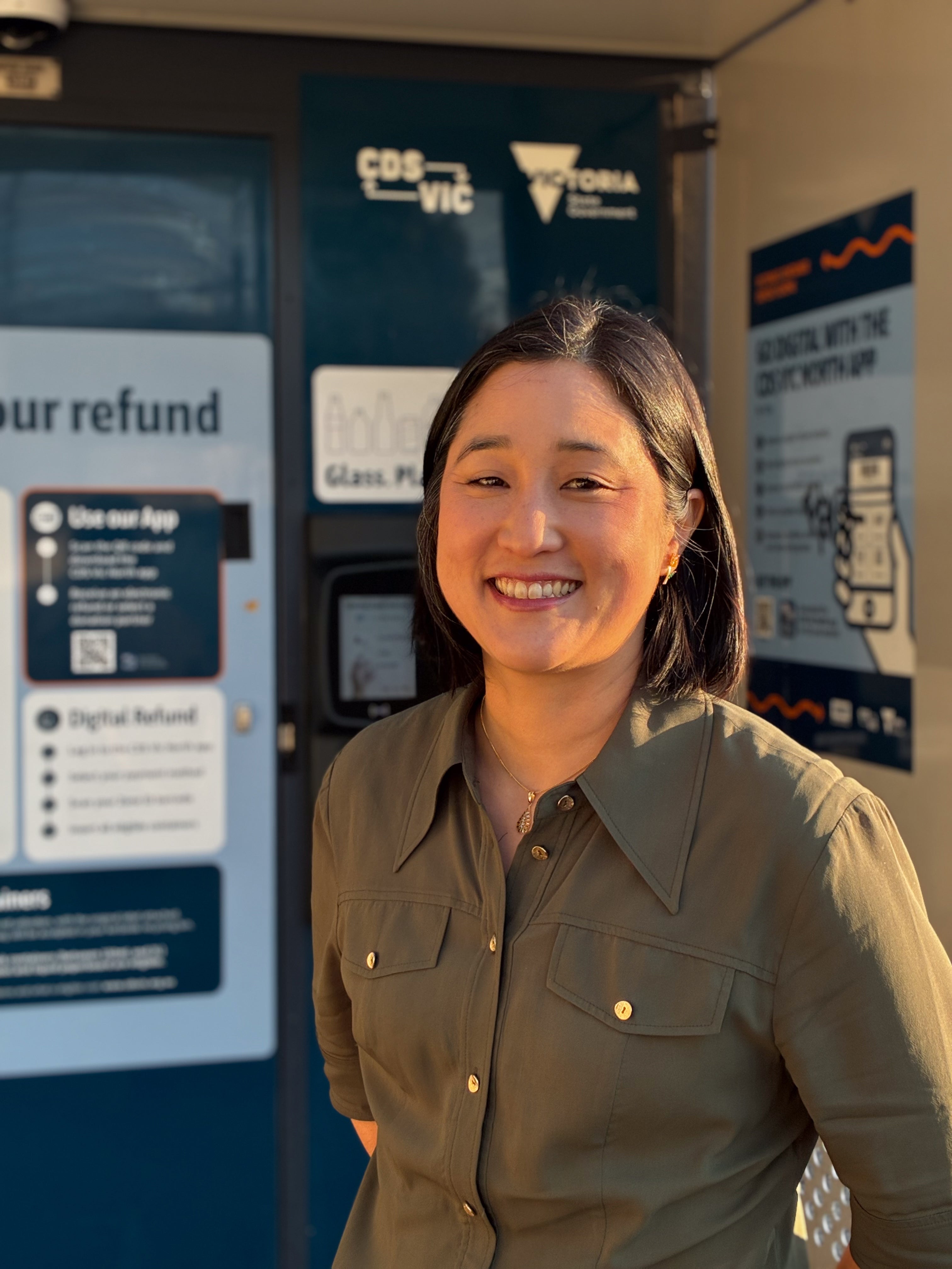 A woman in khaki shirt smiles at camera, she is standing in front of a blue machine