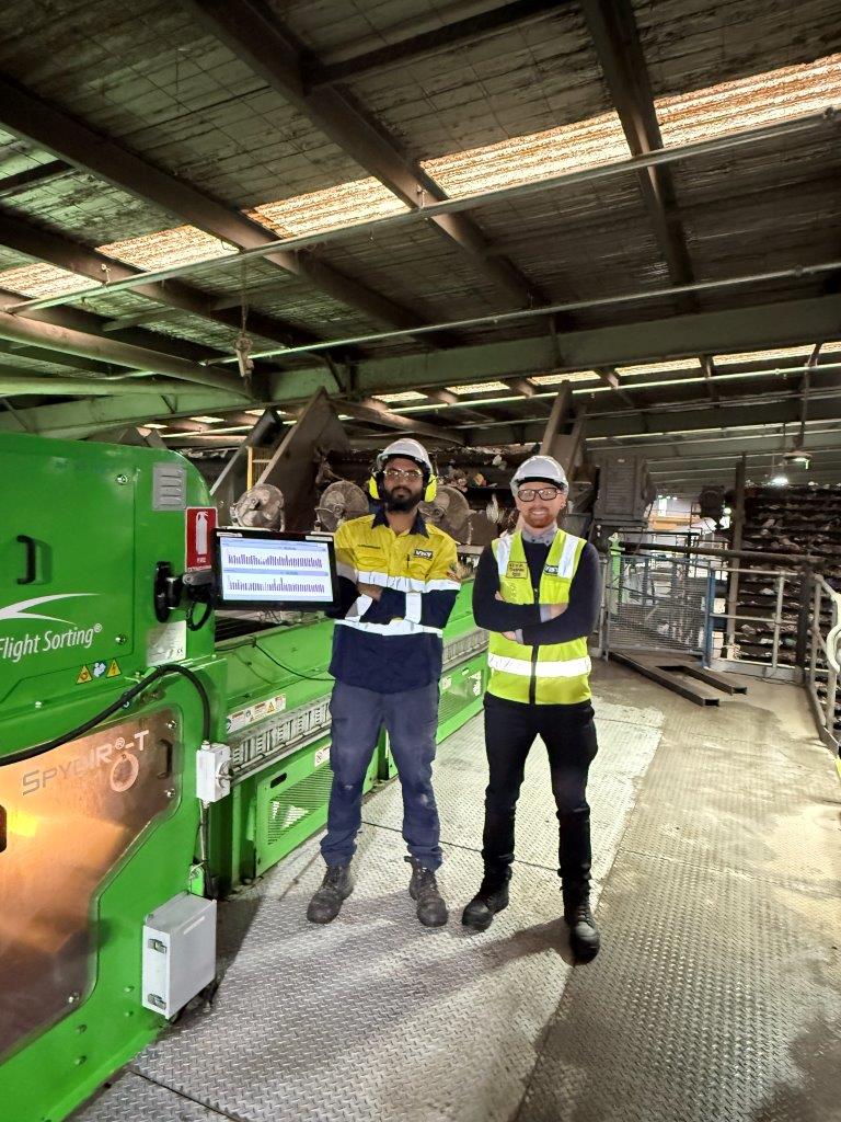 Two men in high visibility safety wear are smiling at the camera with arms folded, they are in a warehouse with equipment behind them