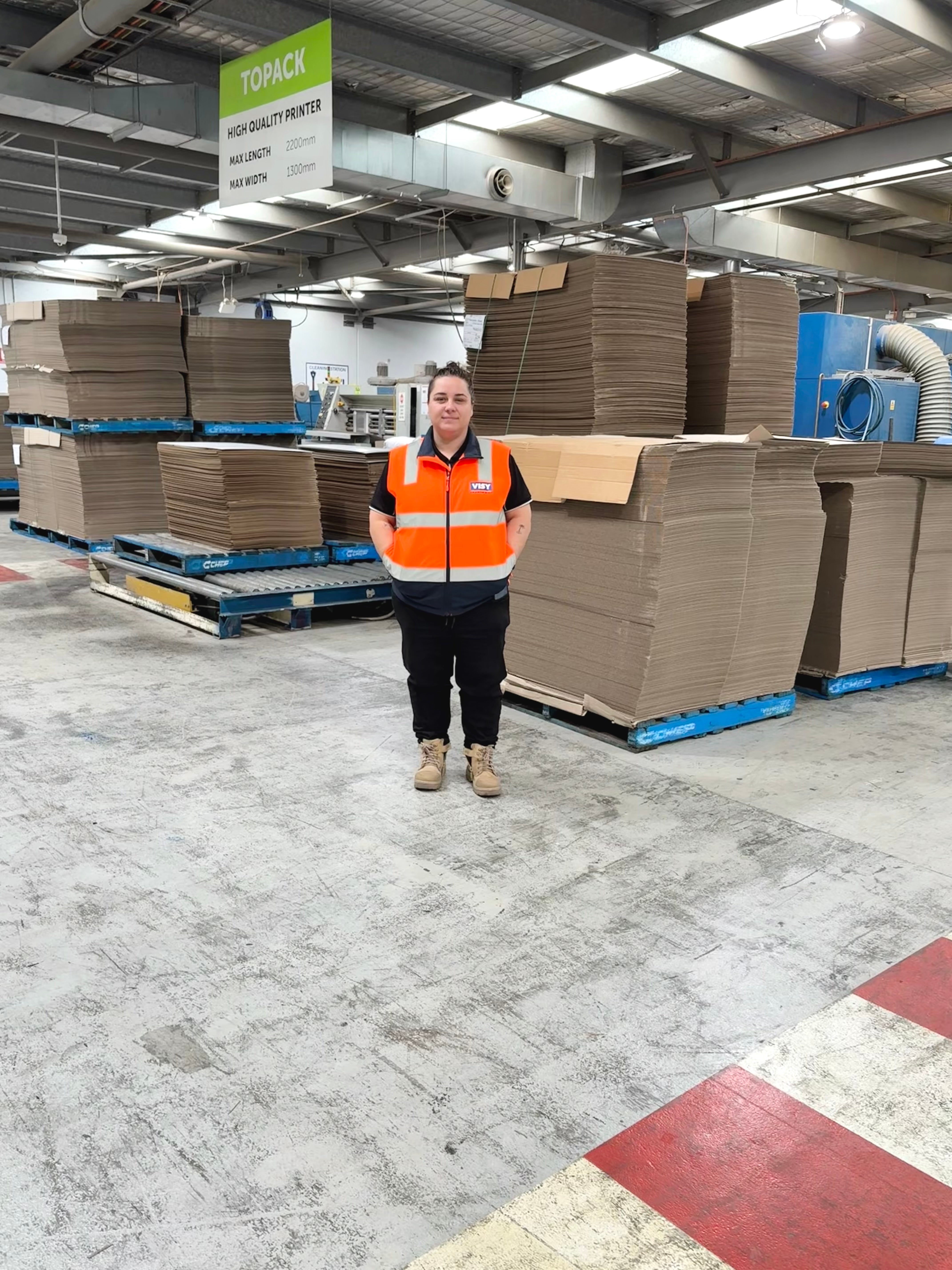A woman wearing an orange high vis safety top smiles at camera, behind her is a concrete area with stacks of brown cardboard