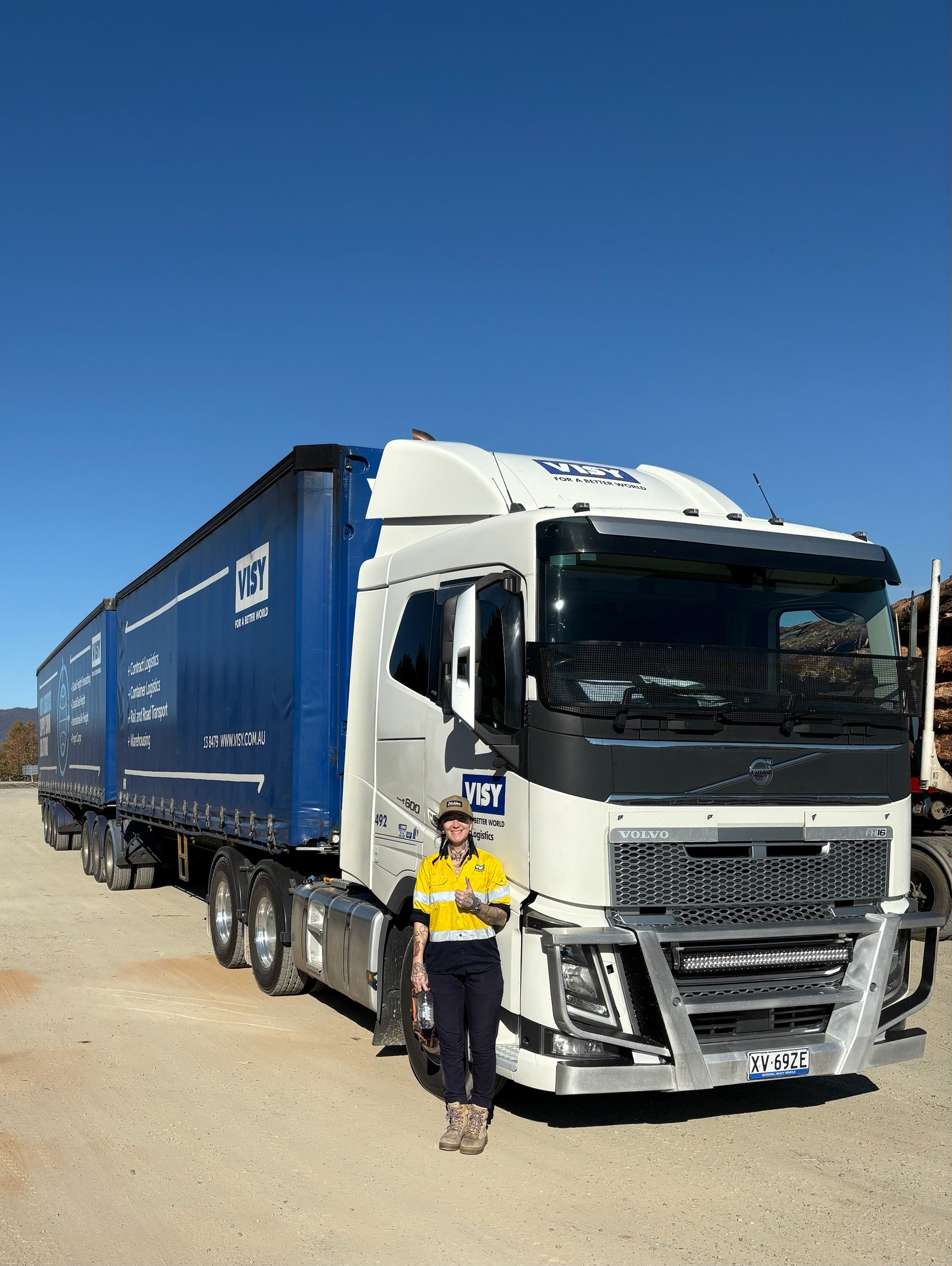 A woman in yellow high visibility work wear smiles with a thumbs up to the camera, she is standing in front of a large blue truck