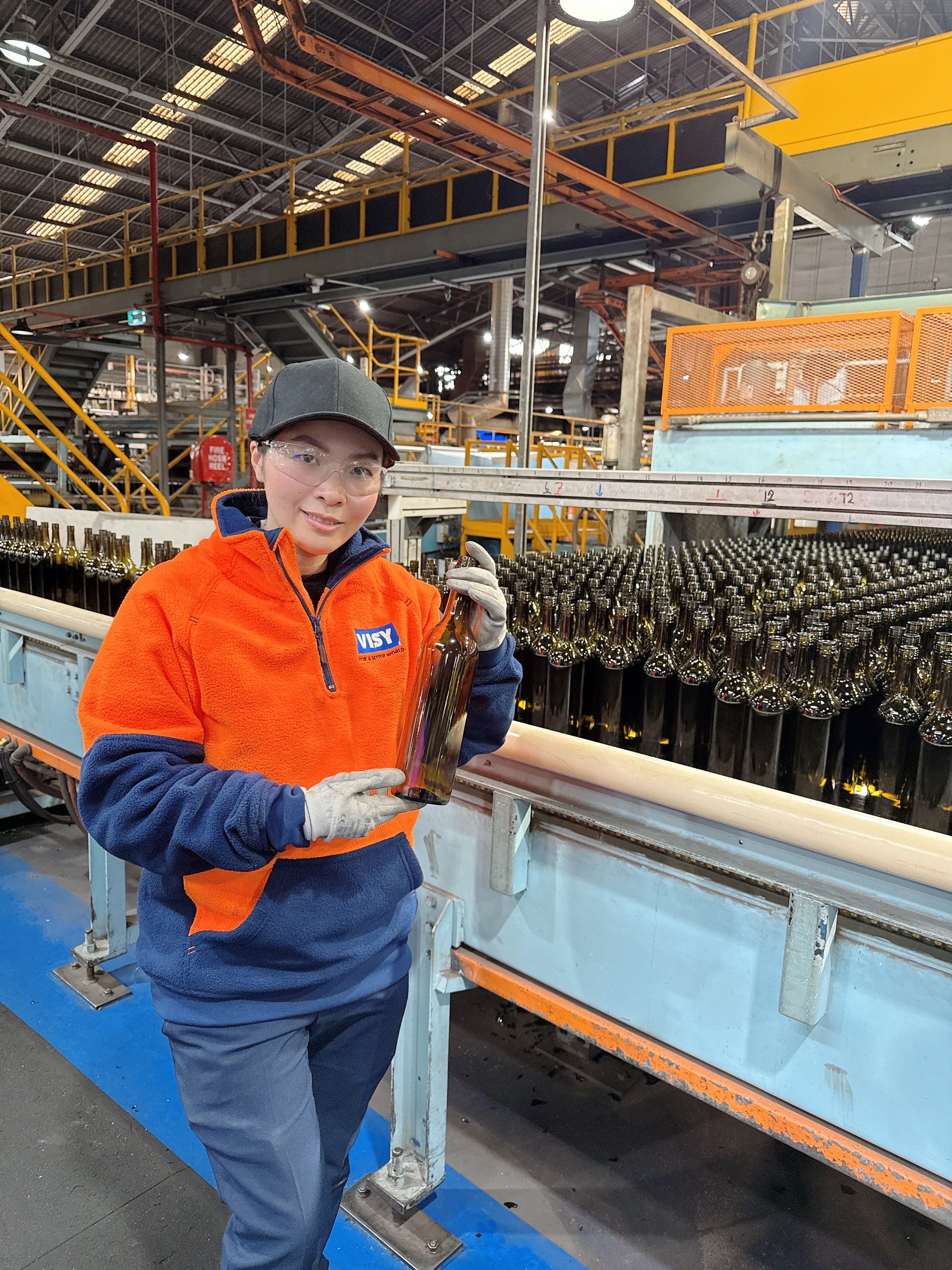 A woman with black cap and safety glasses smiles at camera holding a green wine bottle. She is in a factory with lots of green wine bottles behind her on a conveyor belt. 