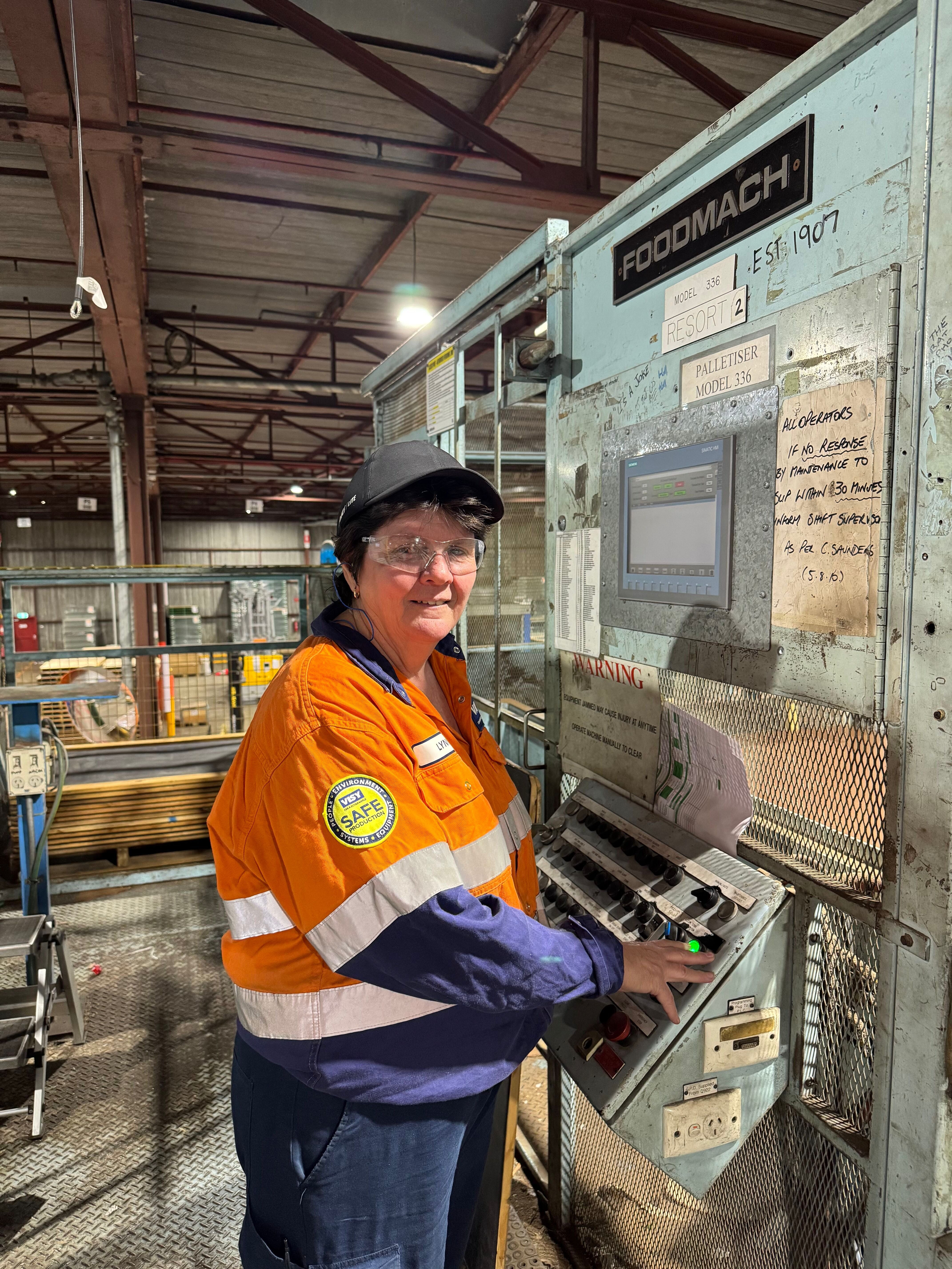 A woman smiles at camera in a black cap, orange and blue high vis safety work wear standing in a factory