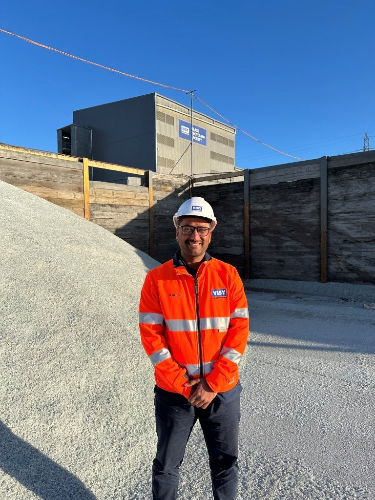 Man smiling at camera in orange high vis work wear with white hard hat. Standing in front of clear recycled glass and a large factory. 