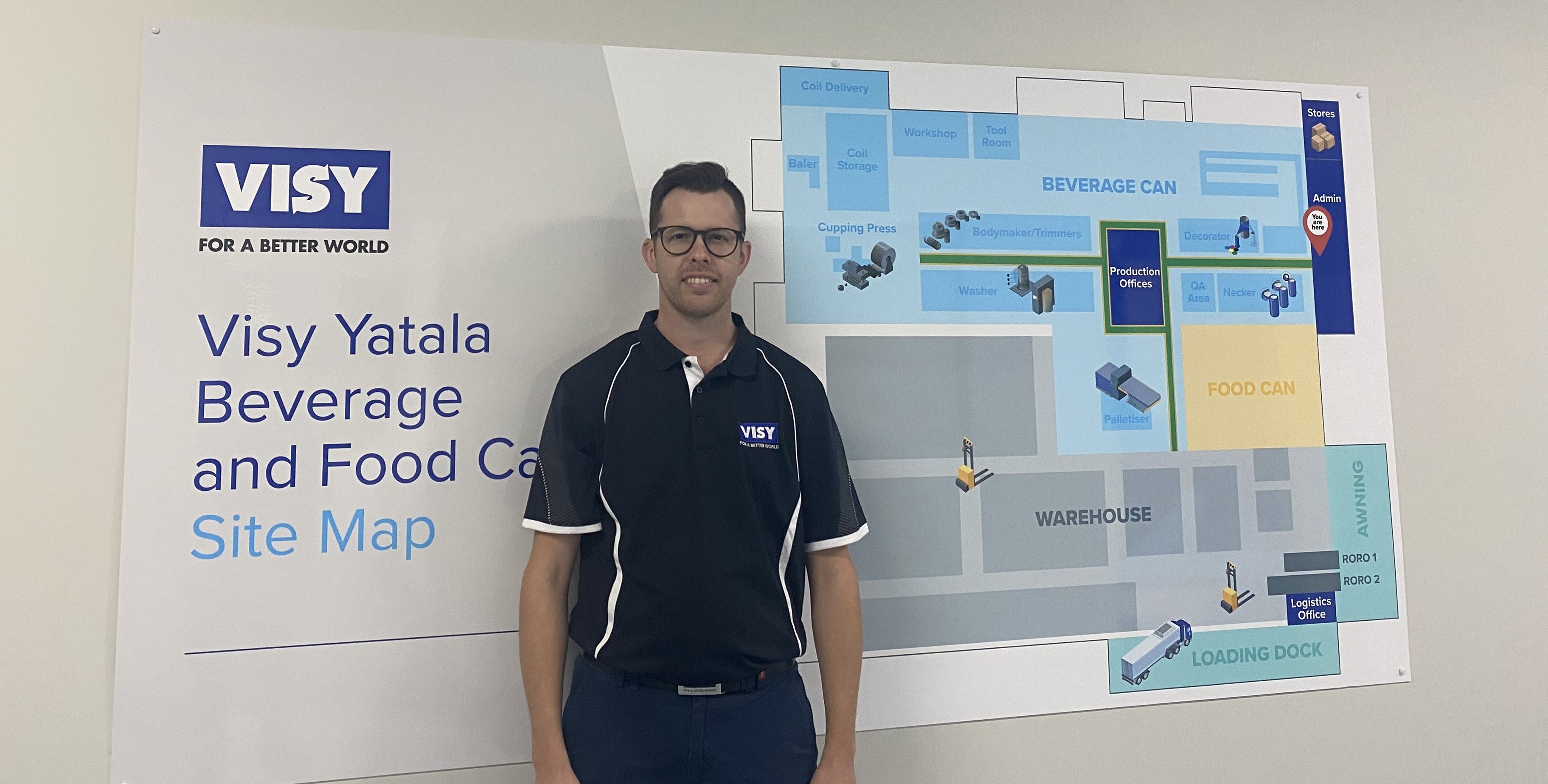 A man smiles at camera in navy blue clothes, he is standing in front of a site map for Visy Yatala beverage and food can