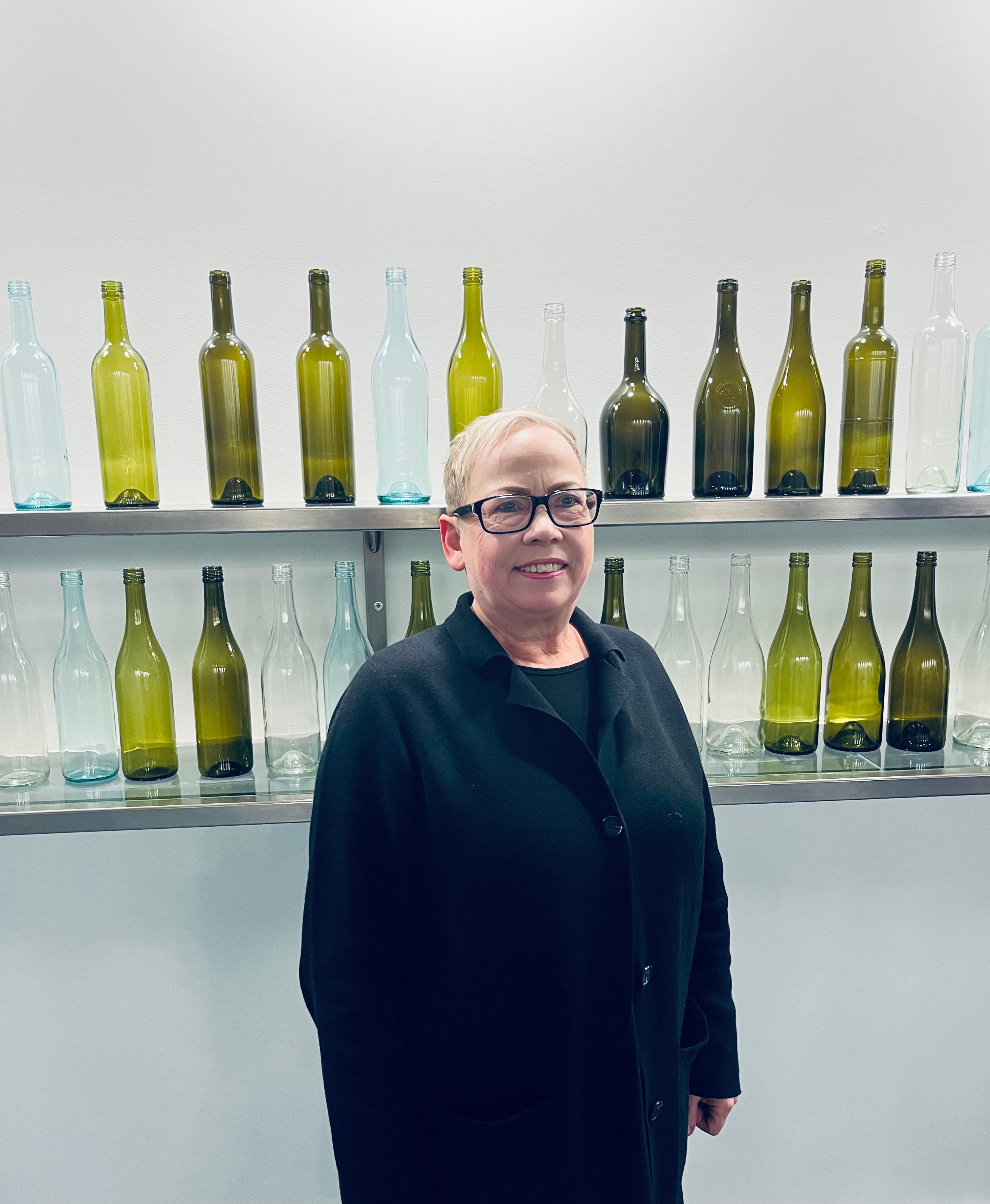 Woman in glasses and dark top smiles at the camera with a wall of glass bottles behind her.