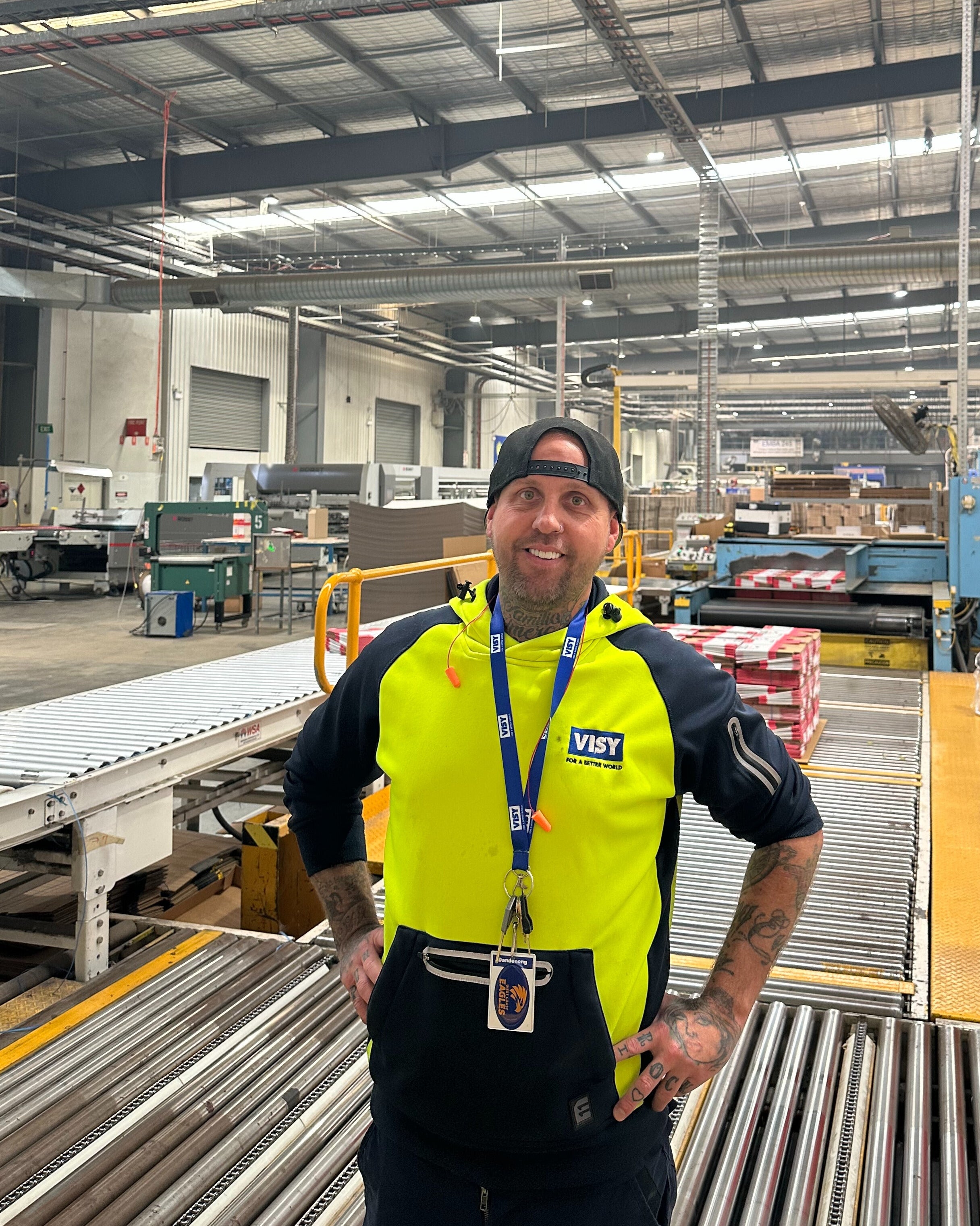 Man in high-vis top and baseball cap stands and smiles at the camera, with cardboard box production behind him.