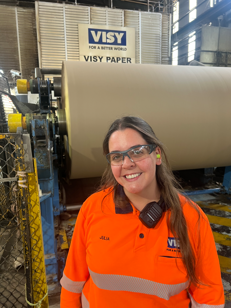 Woman in orange high-vis top smiles at the camera in front of a paper machine.
