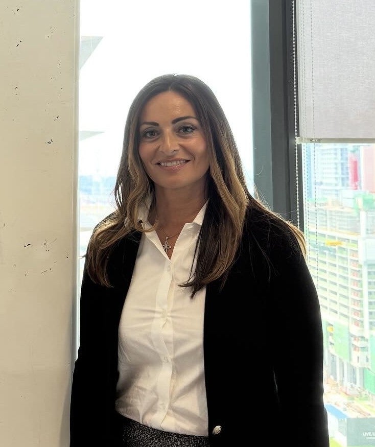 Woman in white shirt and black cardigan smiles at the camera, standing in front of a window.