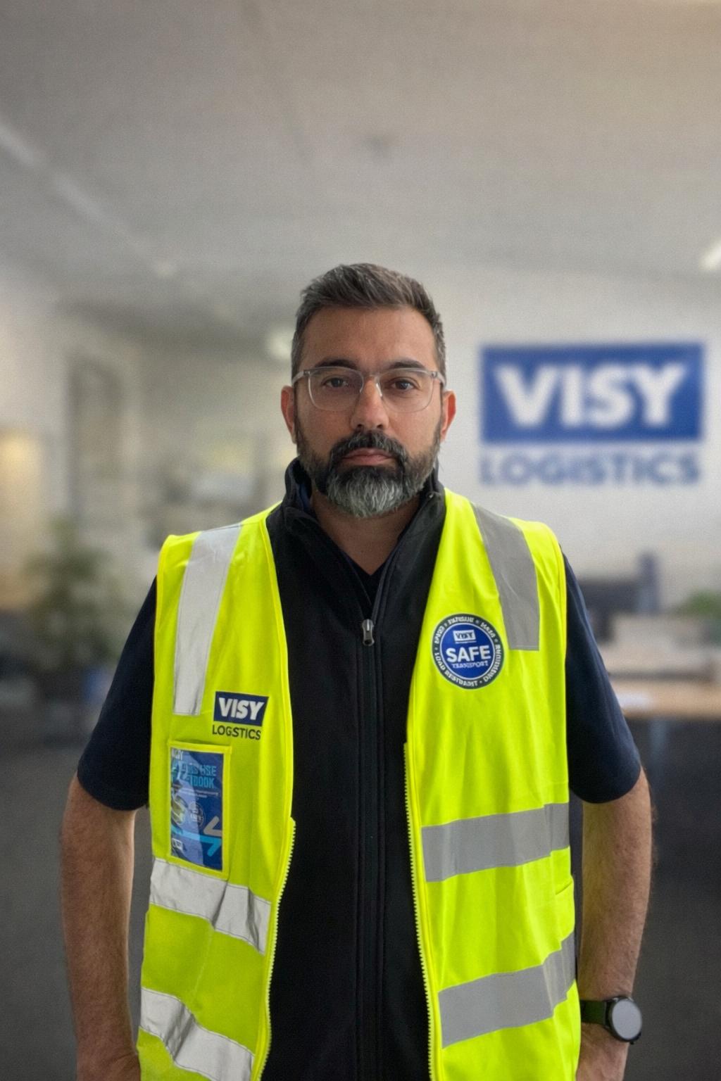 Man wearing yellow high-vis vest and glasses smiles at the camera, in a Visy office.