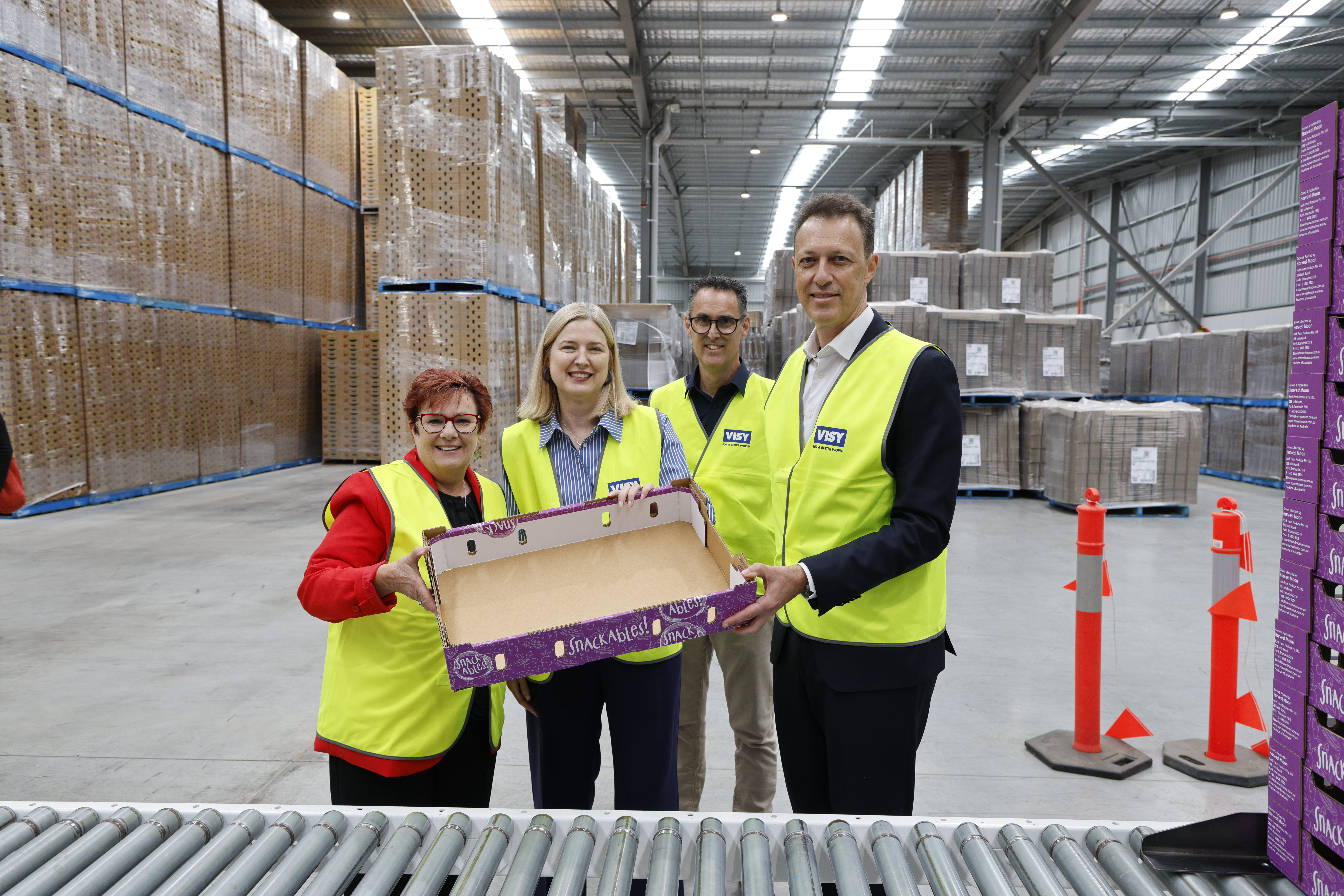 Four people in high visibility safety vests are holding a cardboard produce tray smiling at camera, standing in a warehouse