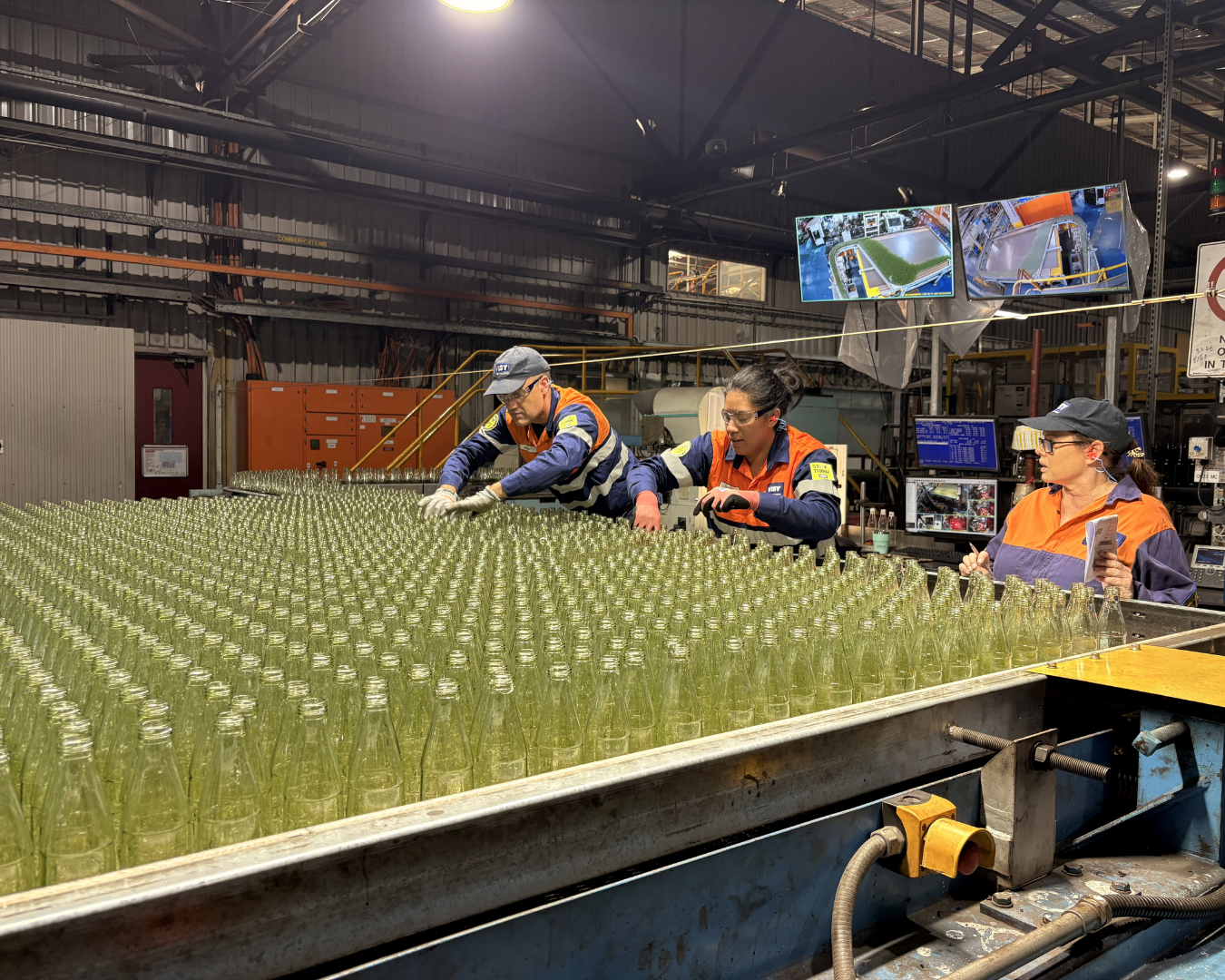 Group of Visy glass employees wearing high-vis, gather at a line of glass bottles as part of their training