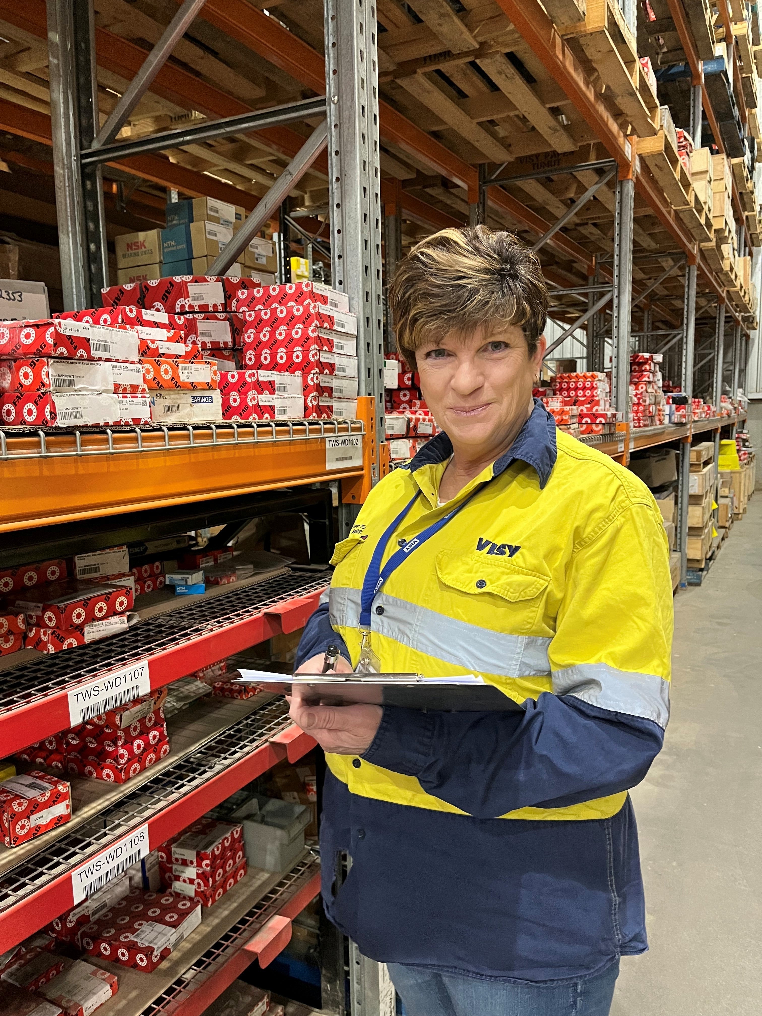 A woman smiles at camera. She is wearing high visibility, safety work wear and holding a clipboard, standing in a warehouse with lots of items on shelves