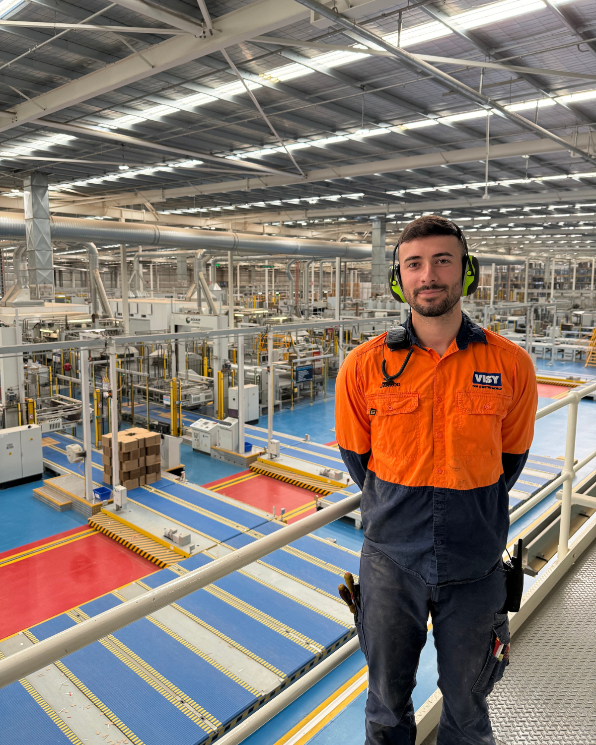 Jarrod, a Visy electrician, is wearing a high-vis orange shirt. He is smiling at the camera in front of a cardboard box facility.
