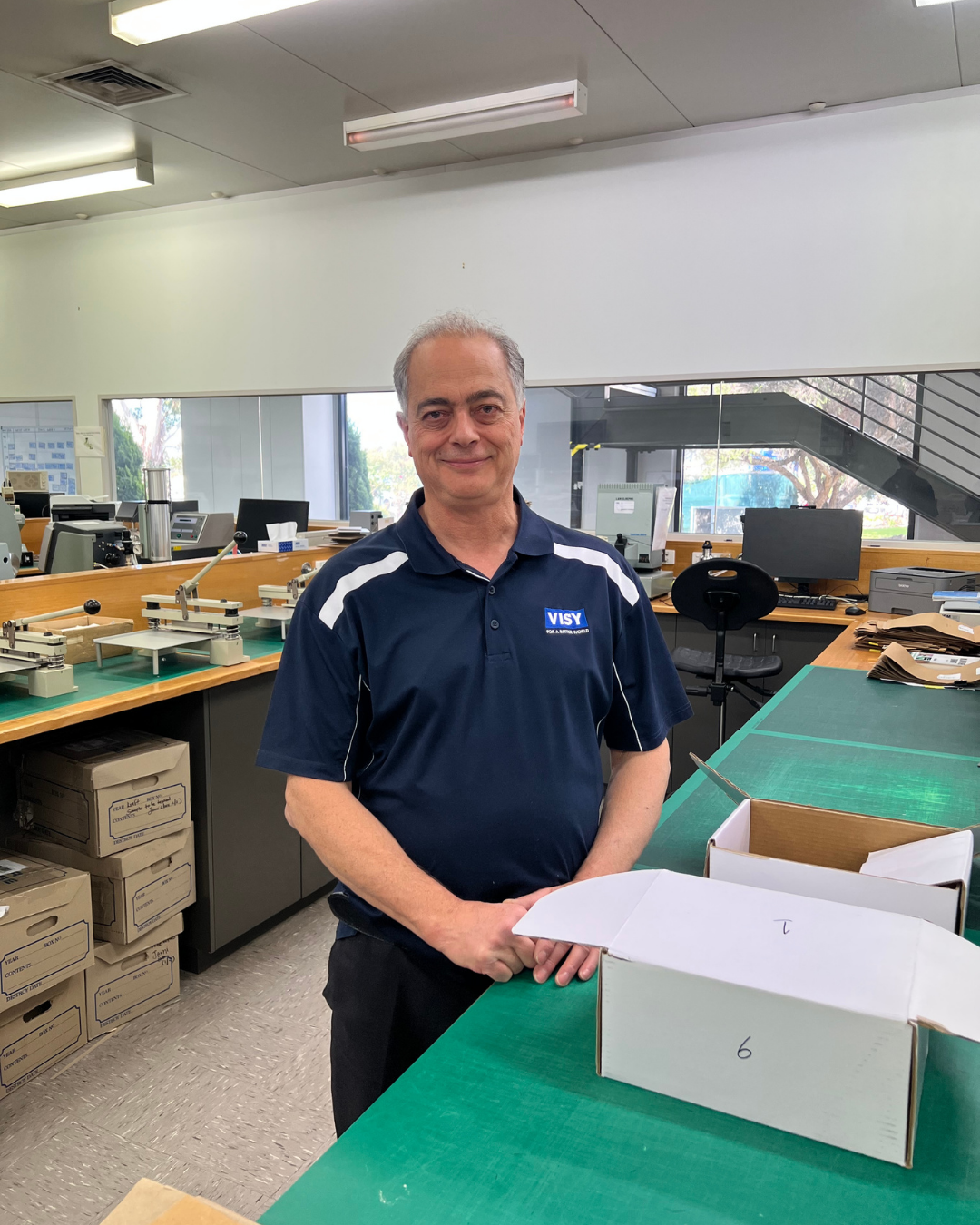 Claudio, a Visy Lab Tech, wears a navy polo shirt with the Visy logo. He stands in a laboratory and smiles.
