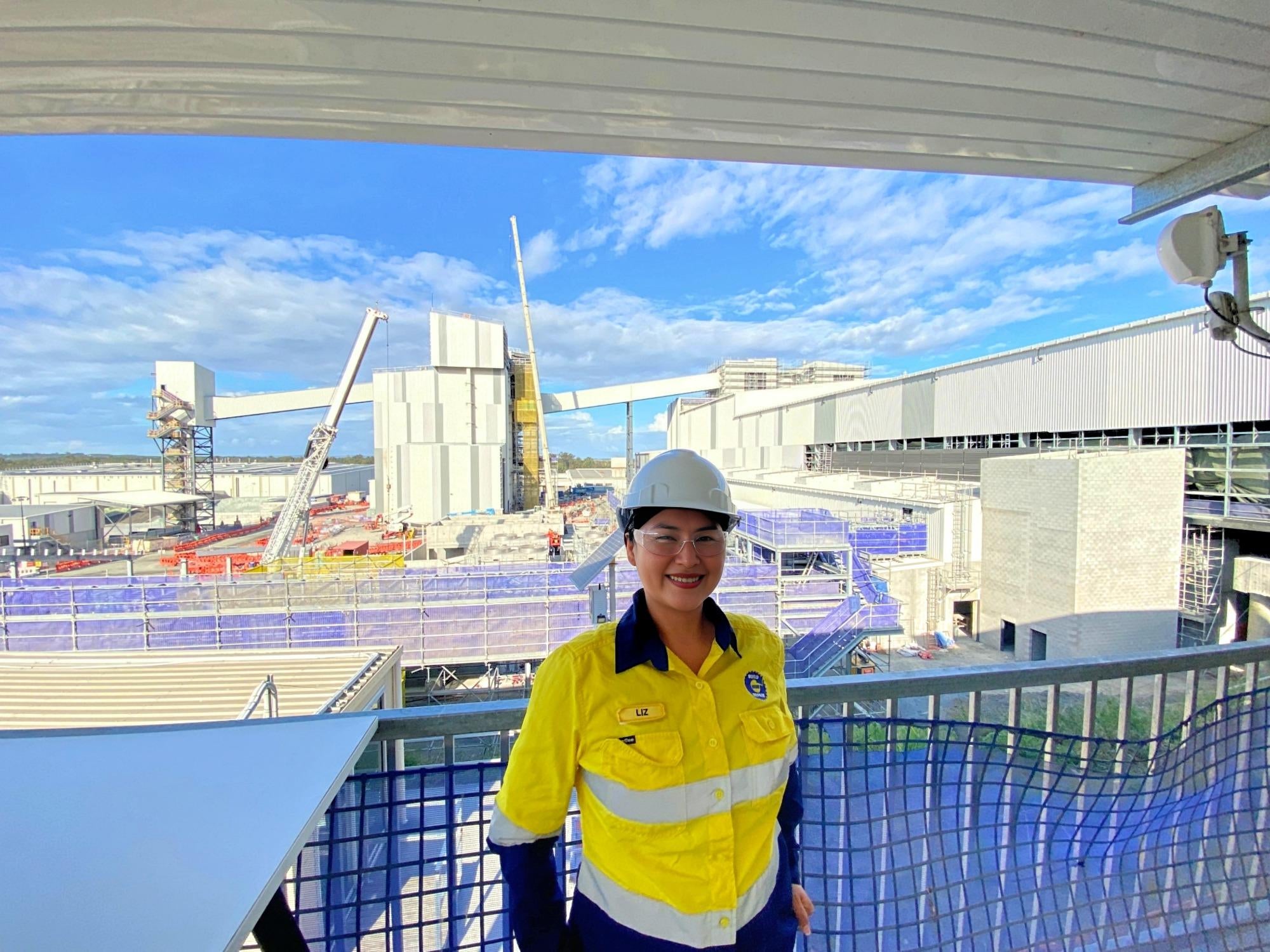 Liz, wearing yellow high-vis, safety goggles and hard hat, smiles in front of construction at Visy Glass Yatala.