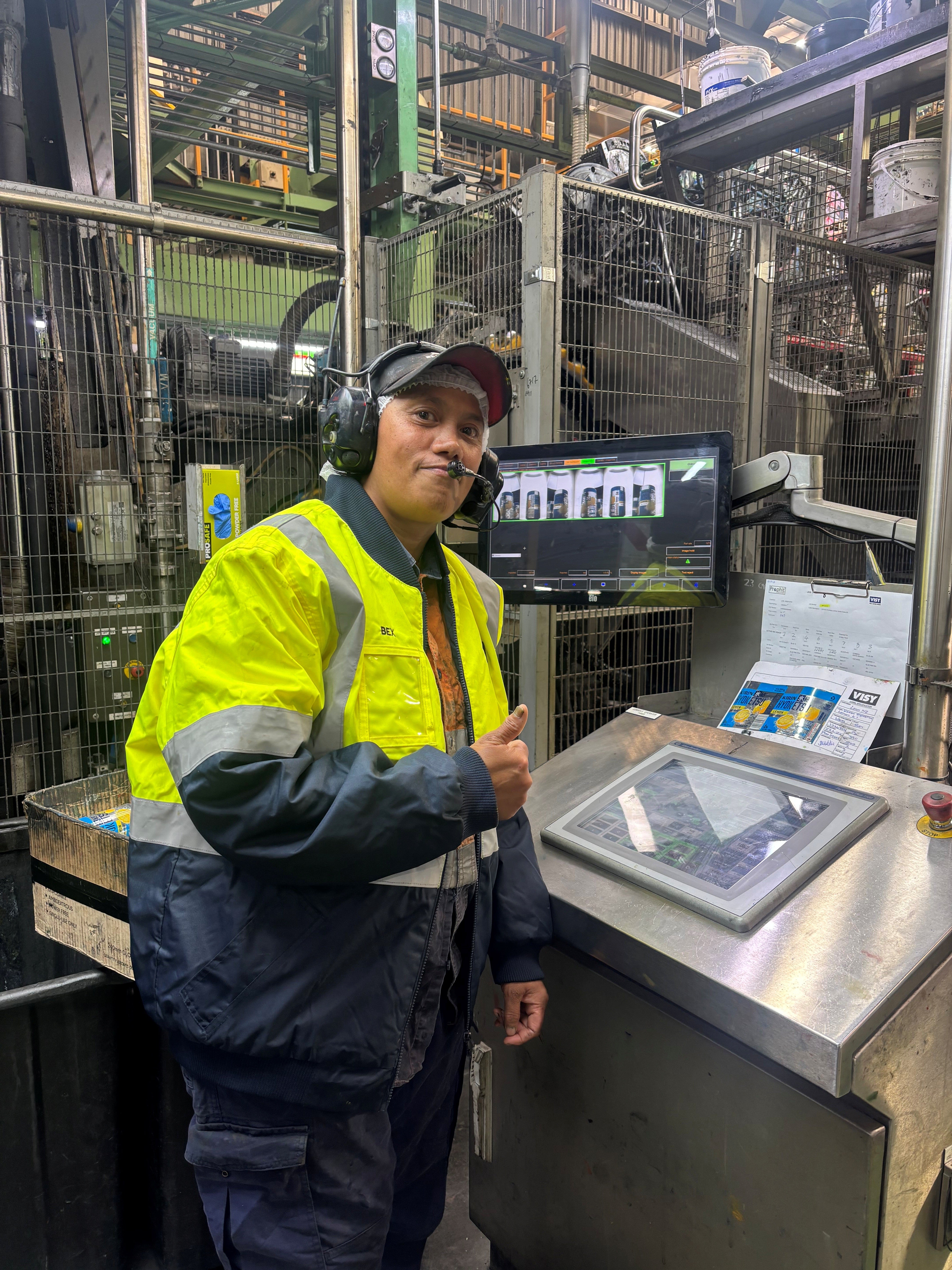 Rebecca wears PPE and smiles at the camera with a thumbs-up. She is standing next to beverage can making equipment.