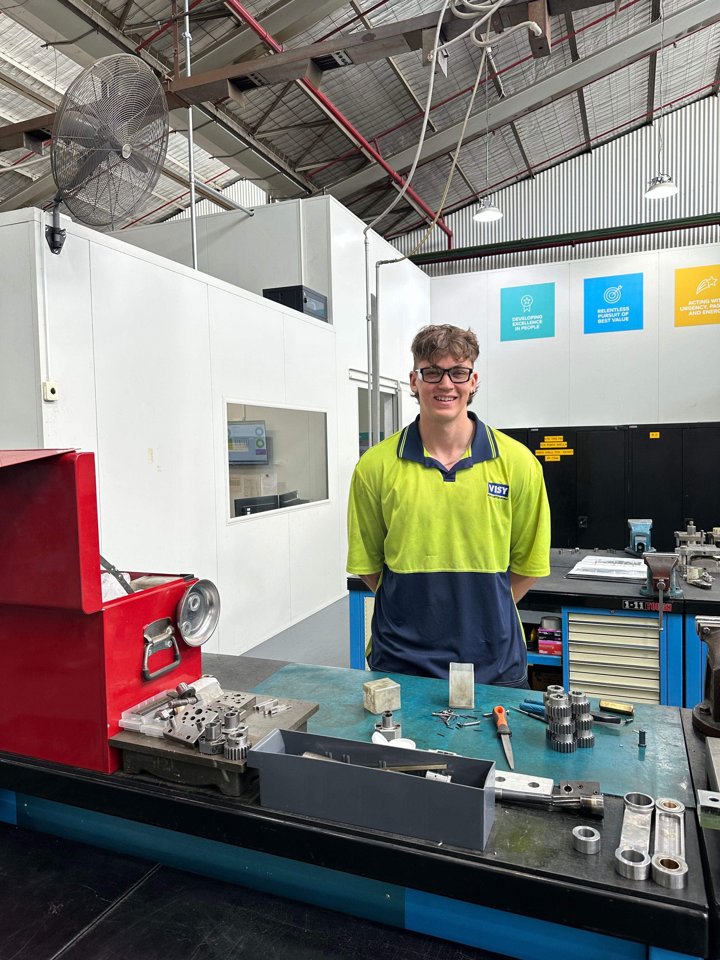 Max wears high-vis and PPE, and smiles at the camera while working in the tool shop