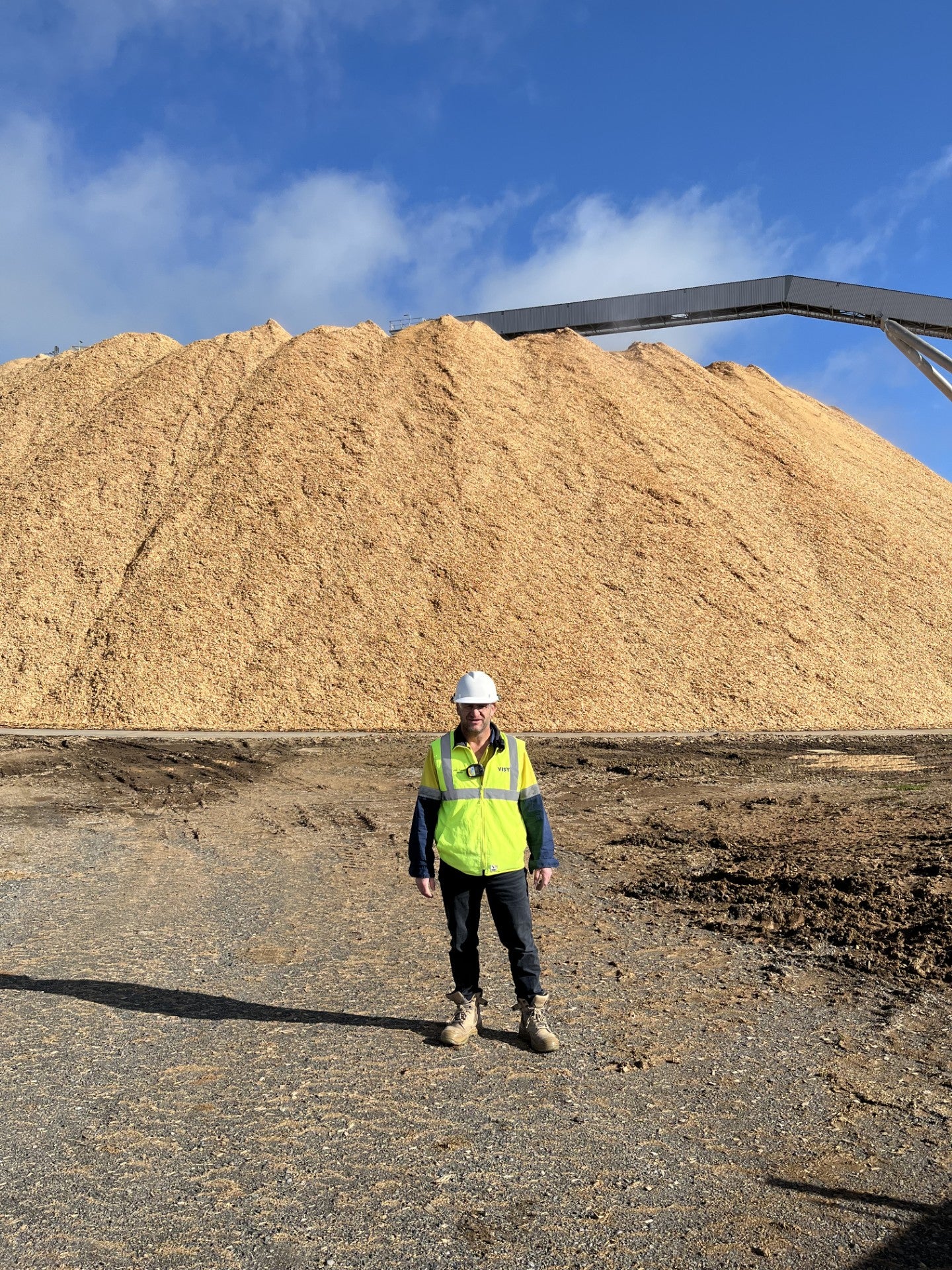 Brad wears PPE and high-vis vest, and stands and smiles in front of a large pile of wood bark..