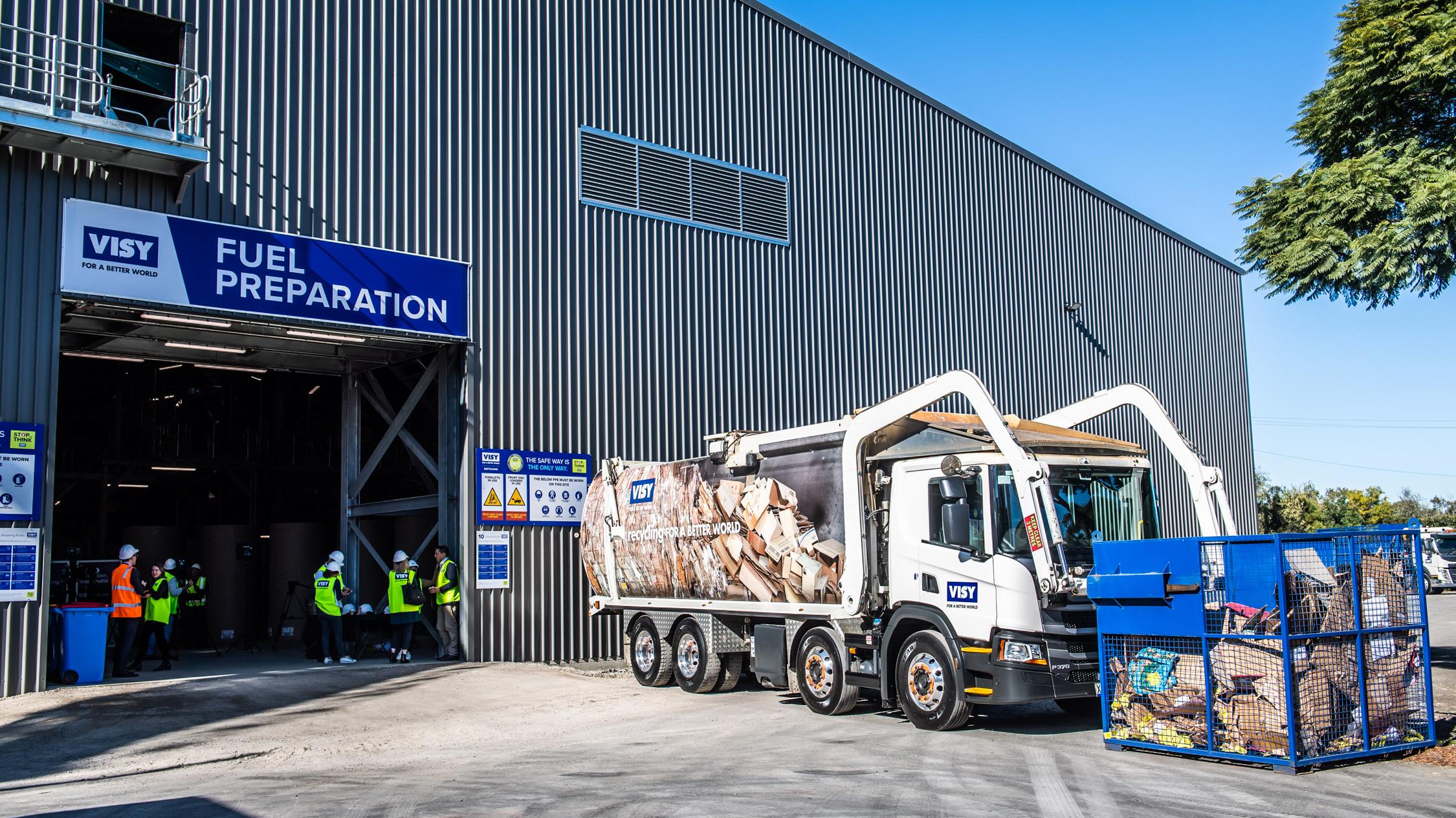 A front loading truck with a bin of cardboard for recycling in front of a shed