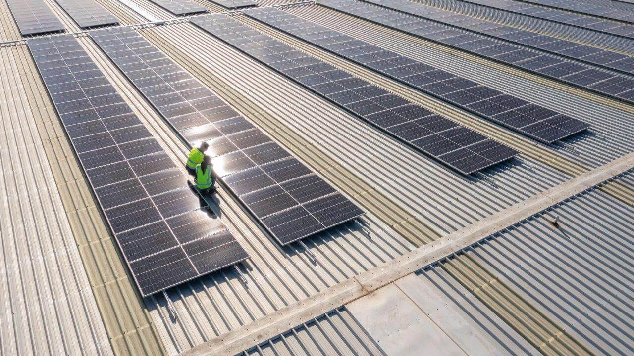 Two people in high visibility safety vests are on a roof looking at solar panels