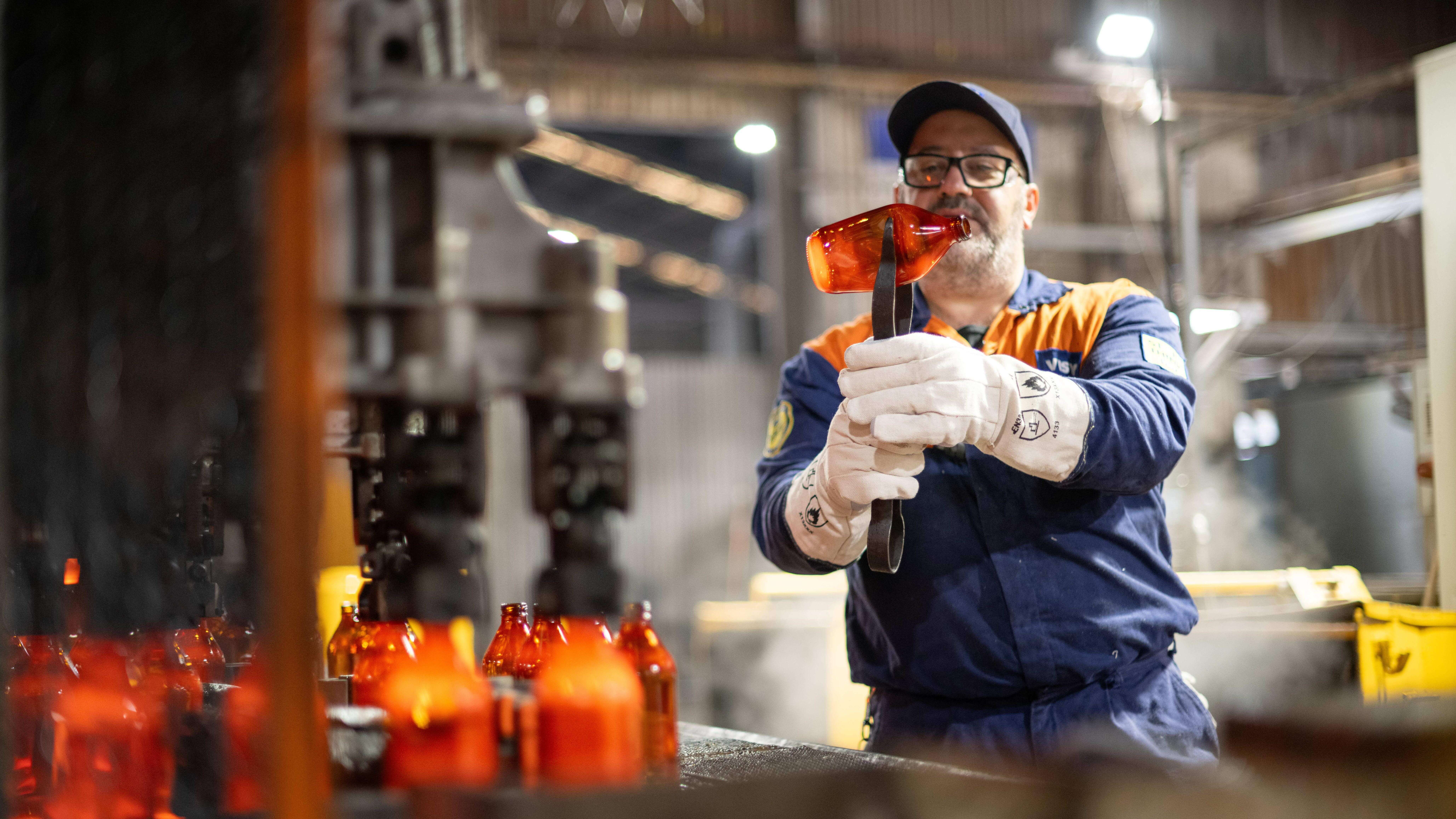 Man in hot end of glass factory holding a glass bottle with tongs