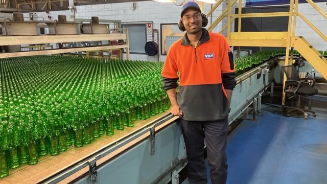A young man in an orange high visibility safety jumper, wearing safety glasses and hard hat is smiling at the camera. He is standing in a factory in front of green glass bottles. 