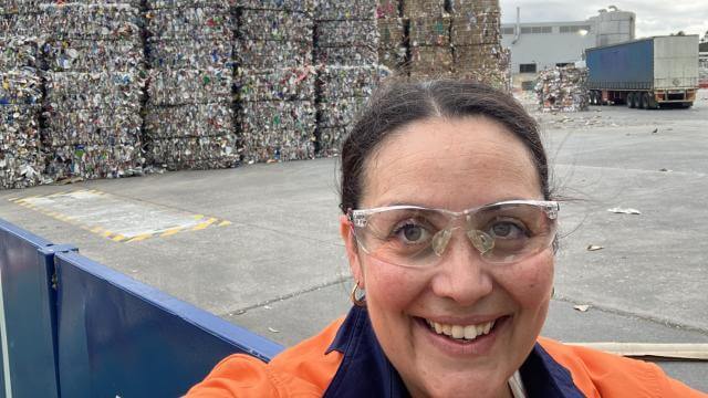 A woman wearing safety glasses and an orange high vis safety top smiles at camera, behind her is a concrete area with bales of recycled paper