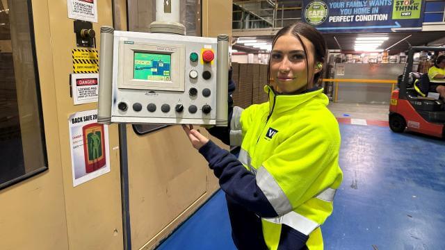 Woman with brown hair in yellow high visibility safety jumper smiles at camera holding a control panel