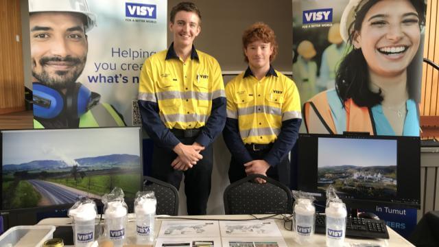 Two young men in high visibility workwear are smiling at the camera, behind a table with Visy branded merchandise 
