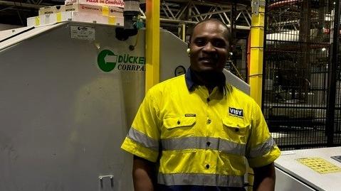 A male employee in a high vis safety shirt smiles at the camera, he is standing in front of machinery