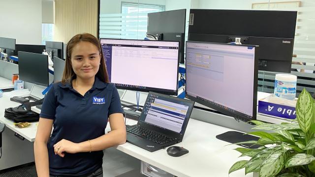 A woman smiles at the camera, wearing a blue Visy-branded polo shirt. She is in an office at a desk with 3 computer screens