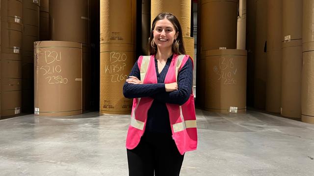 A woman smiles at the camera, she's wearing a pink hi vis safety vest and dark clothes. She's in a warehouse with brown paper reels