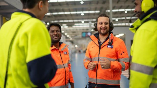 Two men in orange high vis tops are smiling towards the camera talking to two people with their backs to camera