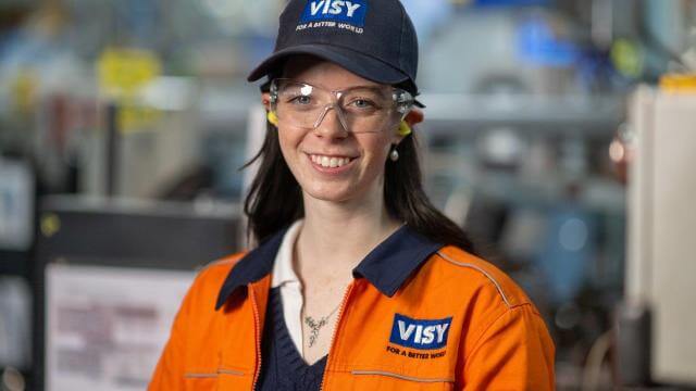 A headshot of a woman in a blue Visy cap, safety glasses and orange high vis top smiles at the camera