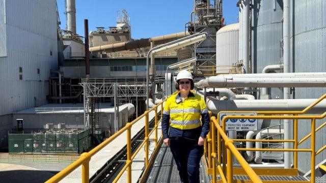 A woman smiles at camera. She is wearing high visibility, safety work wear and a white hard hat. She is surrounding by industrial looking pipes and building. The sky is blue. 