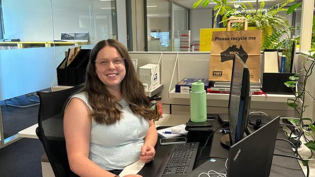 A young woman smiles at the camera, sitting in an office at a computer, she is wearing a light top
