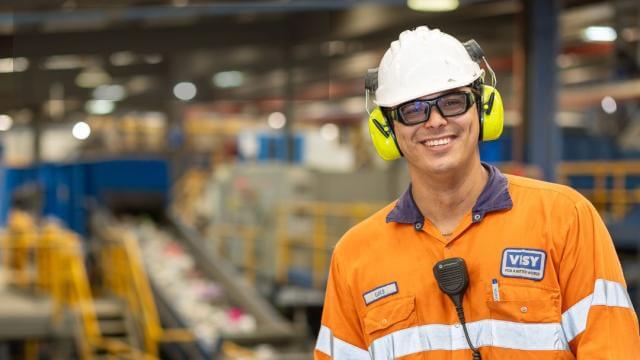 A man in safety gear smiles at the camera, he is in an industrial looking building