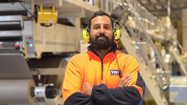 A man in high vis top and ear muffs smiles at camera with arms crossed, he is in an industrial looking building