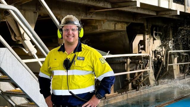 A man smiles at the camera in high visibility work wear, he is in an industrial building with machinery behind him