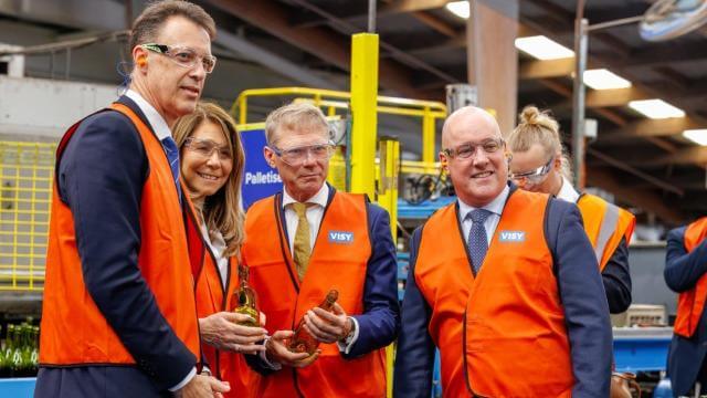 Four people in high visibility safety vests are smiling at the camera in a glass factory
