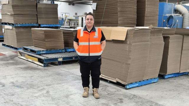 A woman wearing an orange high vis safety top smiles at camera, behind her is a concrete area with stacks of brown cardboard