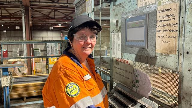 A woman smiles at camera in a black cap, orange and blue high vis safety work wear standing in a factory