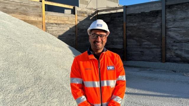 Man smiling at camera in orange high vis work wear with white hard hat. Standing in front of clear recycled glass and a large factory. 