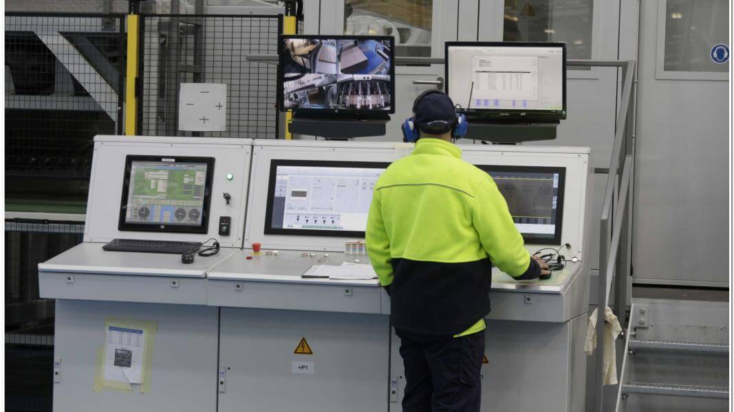 A male employee in high vis and ear muffs works at a computer work station in a factory