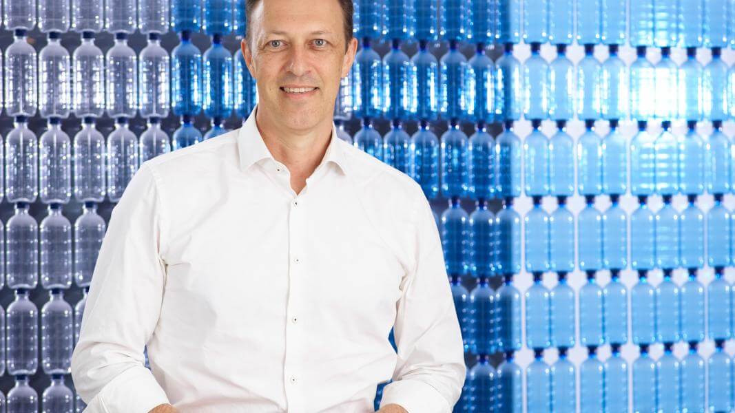 A man in a white business shirt smiles at the camera in front of a background of blue plastic bottles