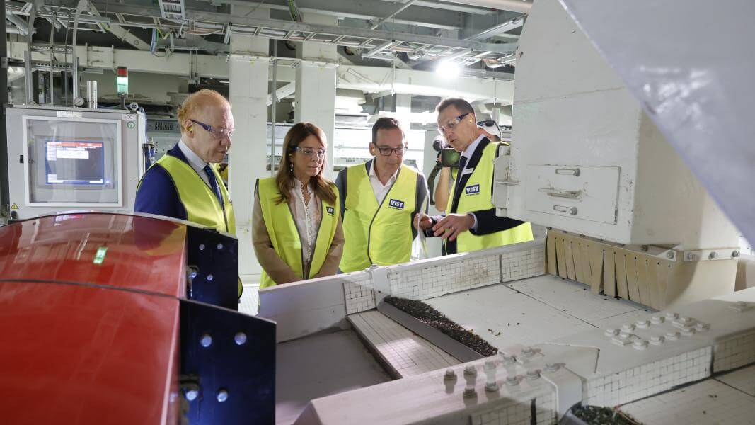 Four people in high visibility yellow safety vests are looking at a glass sorting machine