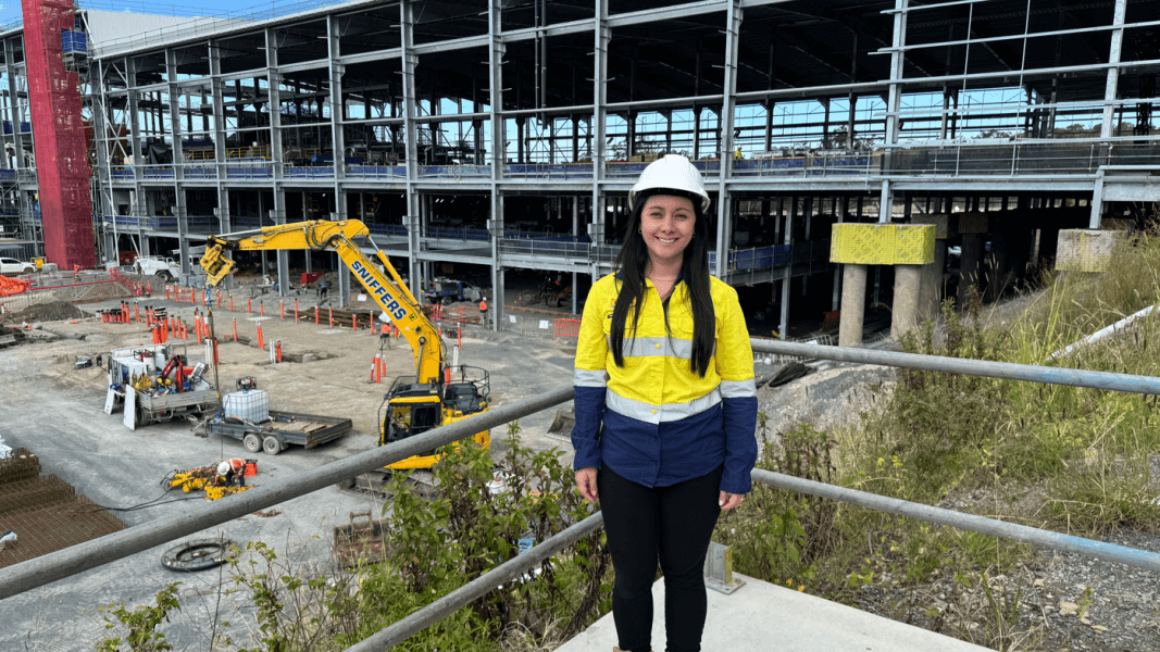 Woman in high-vis top and safety helmet smiles while standing in front of construction site.