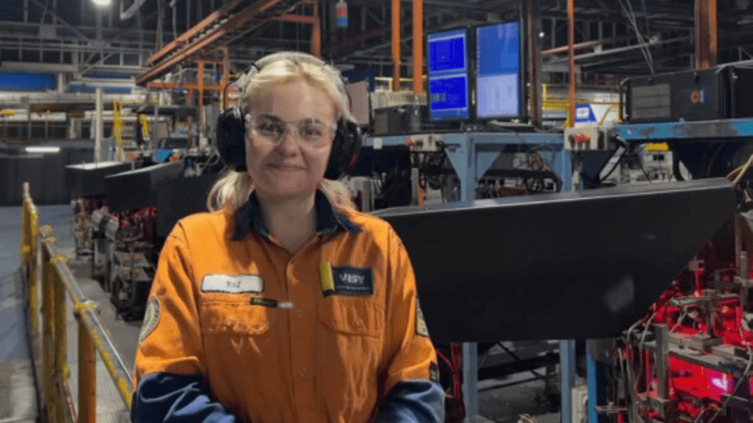 Woman in high-vis top and safety helmet smiles while standing in front of glass machinery.